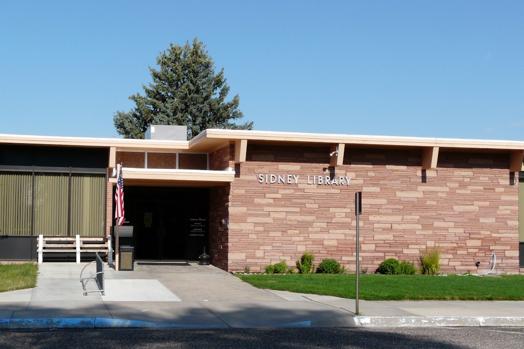 The Sidney Library building: a sandstone brick front with silver lettering and a flag by the entrance